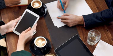 Looking down at a table with people writing, looking at a tablet and drinking coffee.