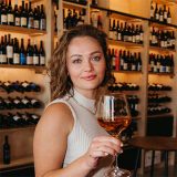 Image of Leigh Fairbrass holding a wine glass in front of racks of bottles of wine.