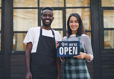 A man and woman standing in front of glass windows holding a sign that says "Yes, we’re open."