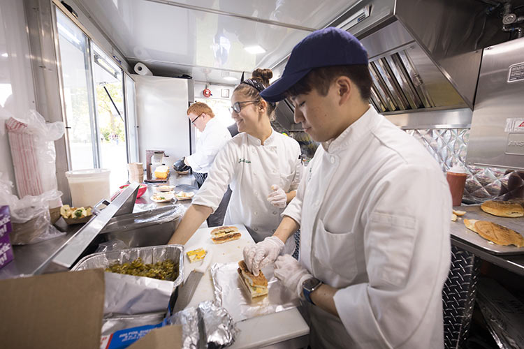 CIA students start a food business by working in a food truck preparing sandwiches in foil sheets.